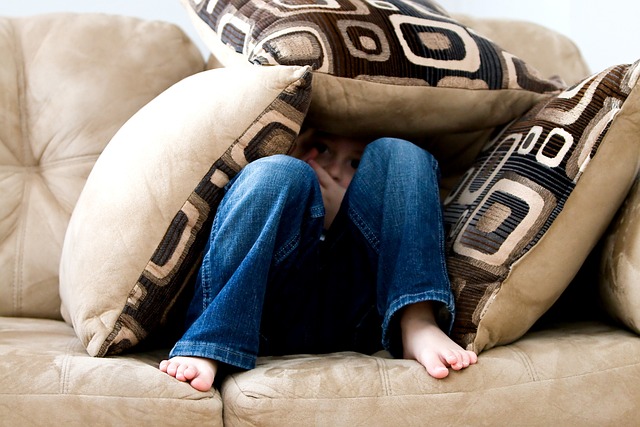 Little boy in blue jeans hiding on a tan suede couch inside a couch cushion fort